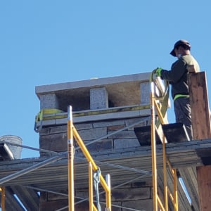 Worker on scaffolding installing stone chimney top with clear blue sky in background, showcasing crane services in construction project.