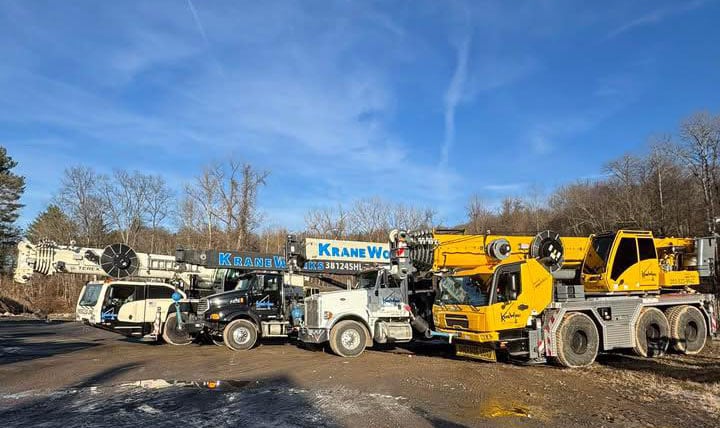 Kraneworks crane fleet featuring all-terrain boom cranes, boom trucks, and equipment in a construction yard under a clear blue sky.