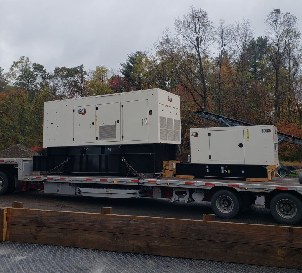 Heavy machinery transport featuring two CAT generators on a flatbed truck, surrounded by autumn foliage, illustrating KraneWorks Inc.'s equipment hauling services.