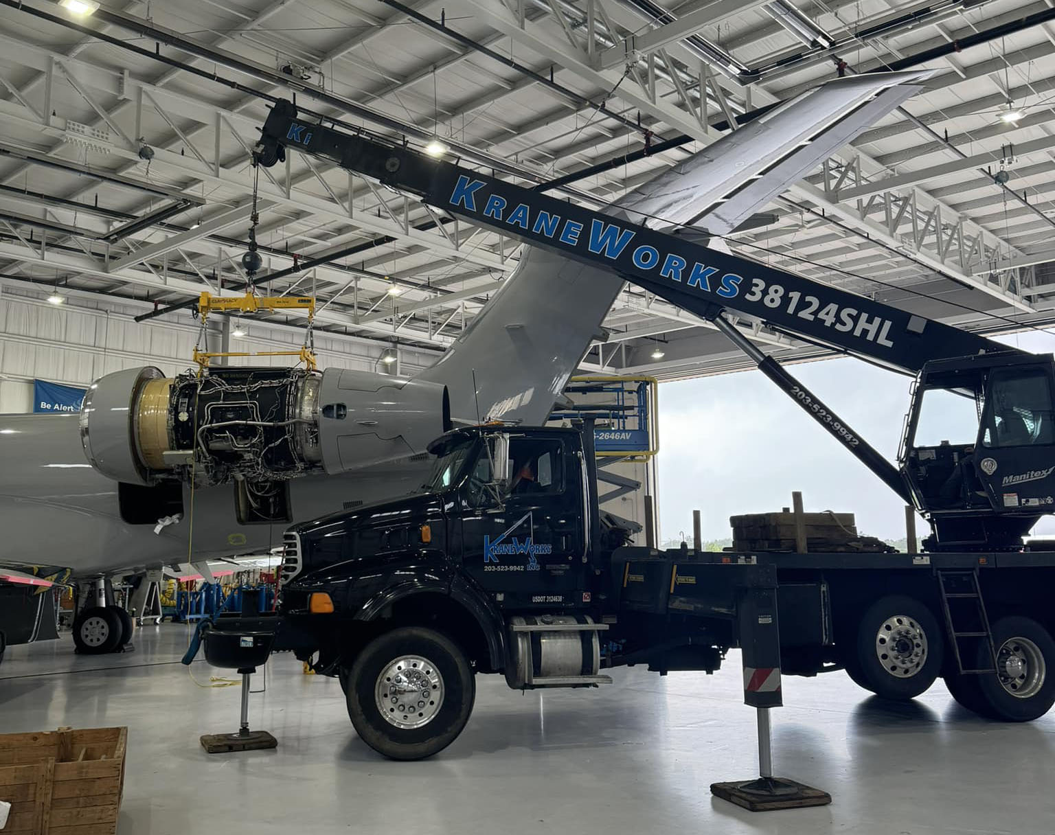 Manitex 38124SHL boom truck lifting aircraft engine inside a hangar, showcasing KraneWorks branding and hydraulic capabilities for residential and commercial lifting tasks.