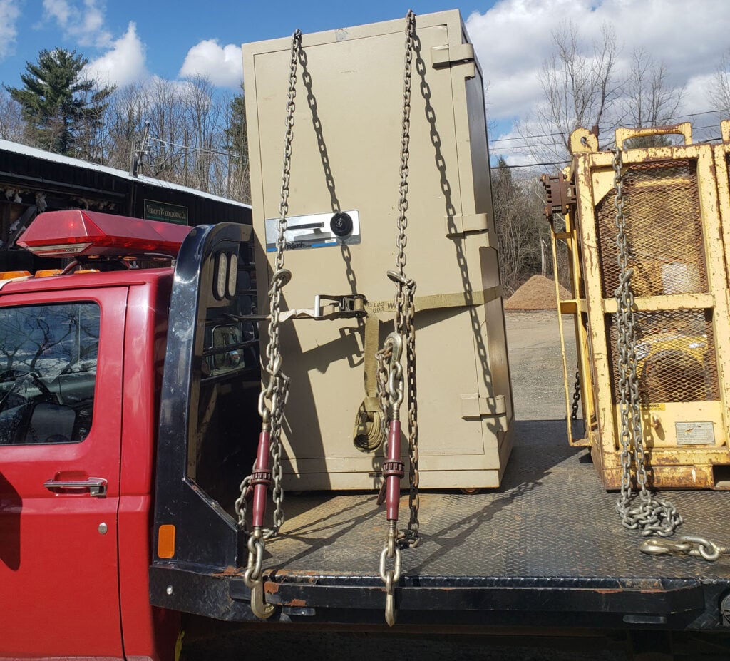 Heavy equipment secured on a red truck's flatbed, featuring a large, beige unit with chains and straps, illustrating KraneWorks' transportation and hauling services.