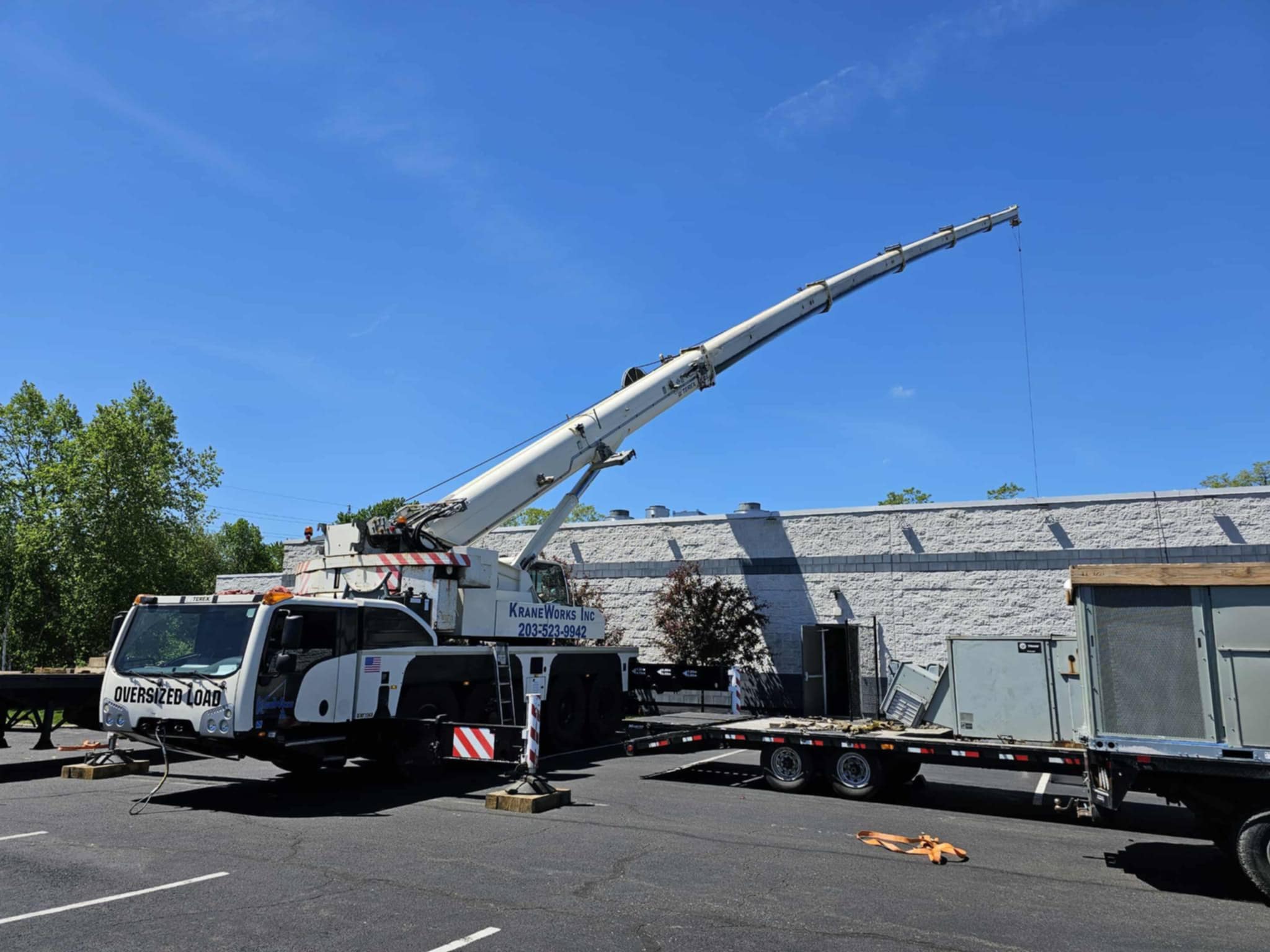 All-terrain boom truck from KraneWorks Inc. with extended boom, lifting equipment from trailer, and "Oversized Load" signage, set against a clear blue sky.